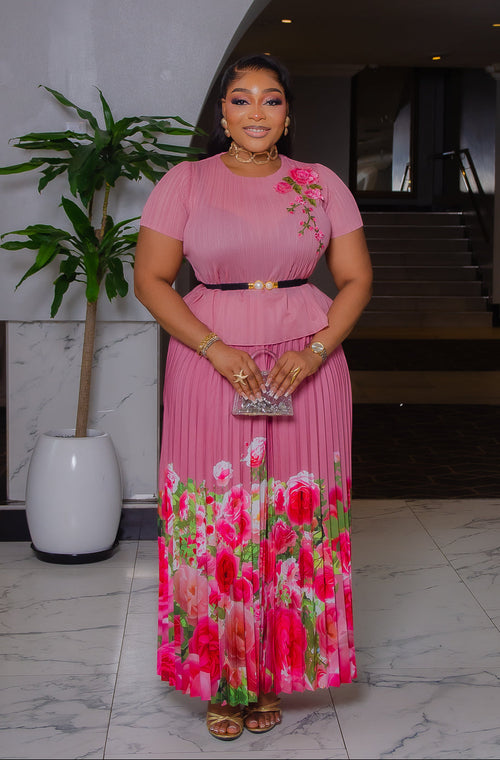 Model wearing Blossom Beauty mauve pink Miyake pleated two-piece set featuring a floral peplum top, pearl buckle belt, and flowing pink rose-print maxi skirt.