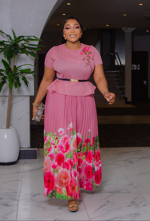 Model wearing Blossom Beauty mauve pink Miyake pleated two-piece set featuring a floral peplum top, pearl buckle belt, and flowing pink rose-print maxi skirt.
