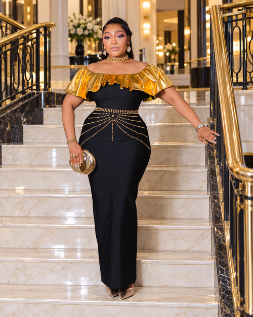 Black and gold off-shoulder bandage maxi dress with metallic gold ruffle neckline and decorative gold beadwork at the waist.
